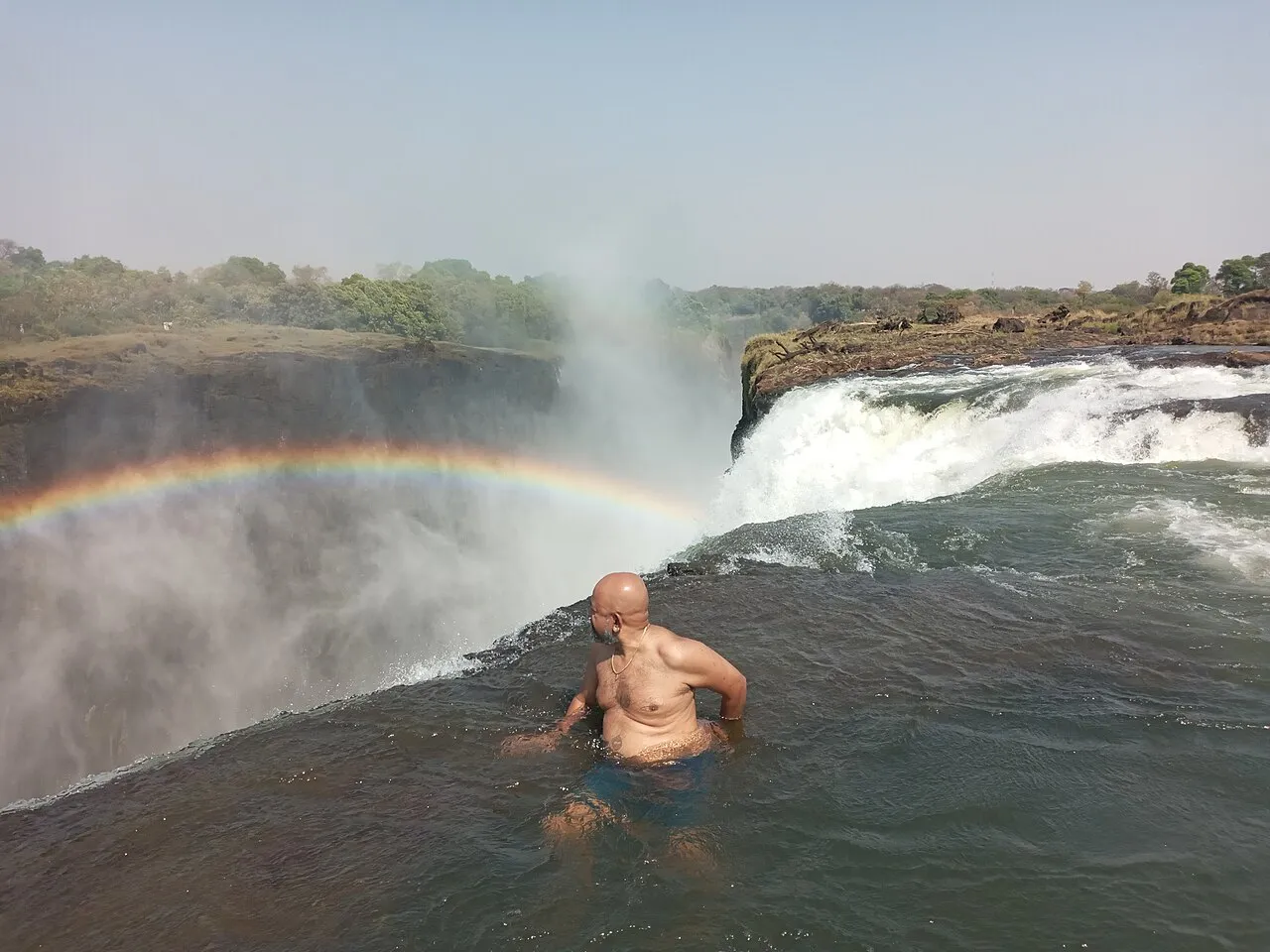 A visitor sits at the rock lip of Devil's Pool on Livingstone Island, with spray and a rainbow rising from Victoria Falls below.