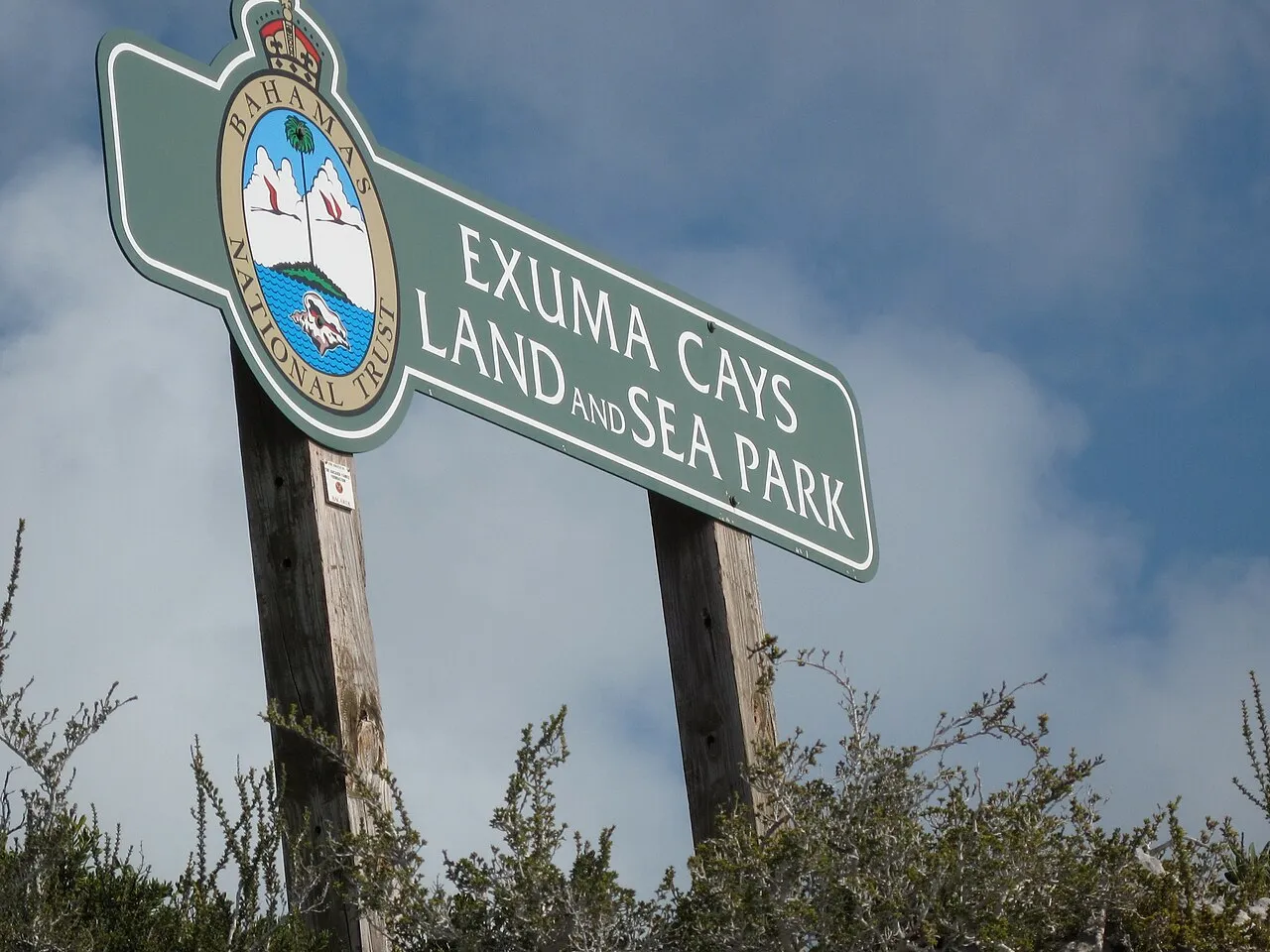 A wooden trailside sign reading "Exuma Cays Land and Sea Park" with the Bahamas National Trust crest, set against sky and scrub bushes.