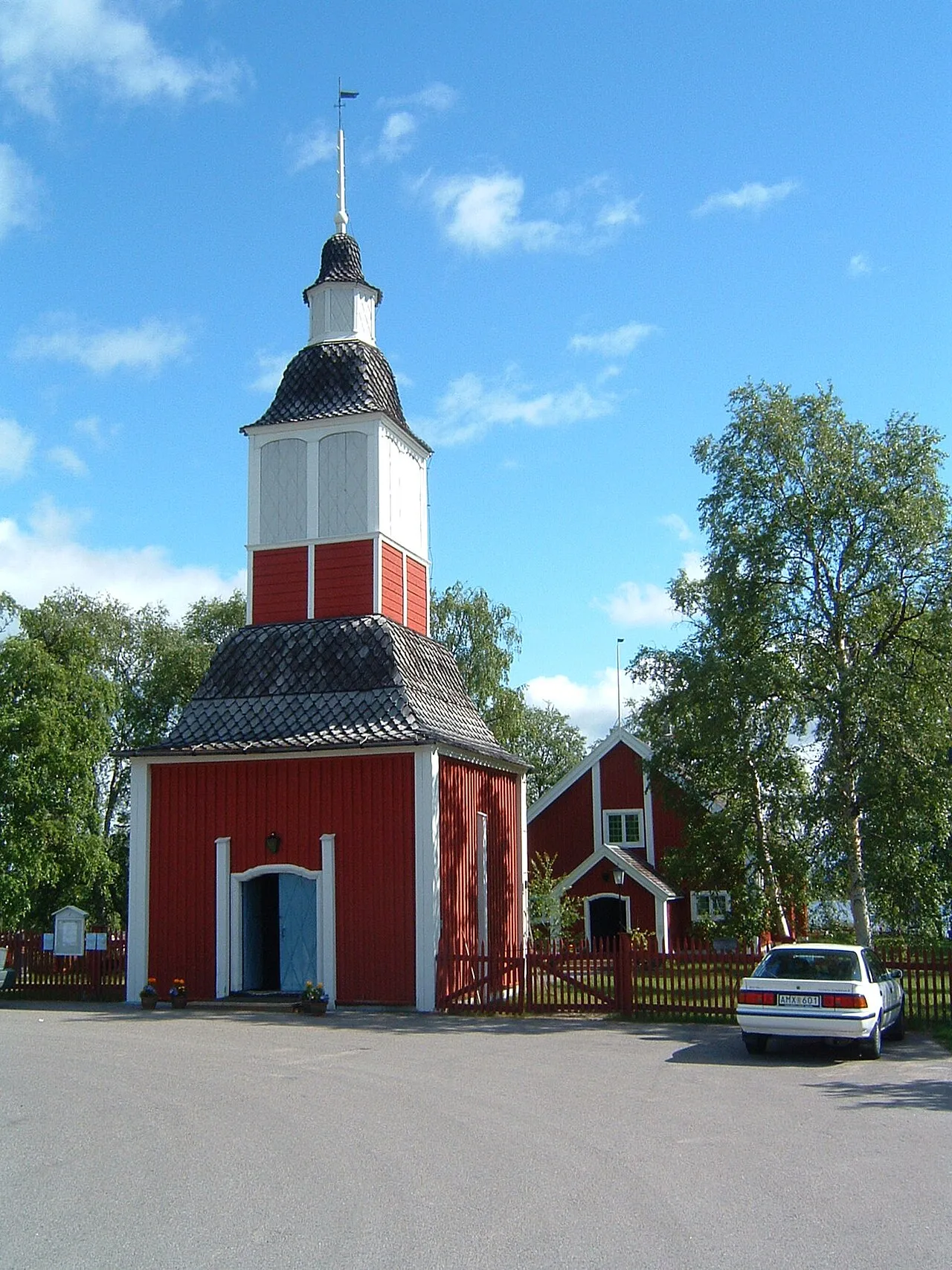 The red wooden bell tower and church of Jukkasjärvi under a blue summer sky, with birches beside the churchyard fence
