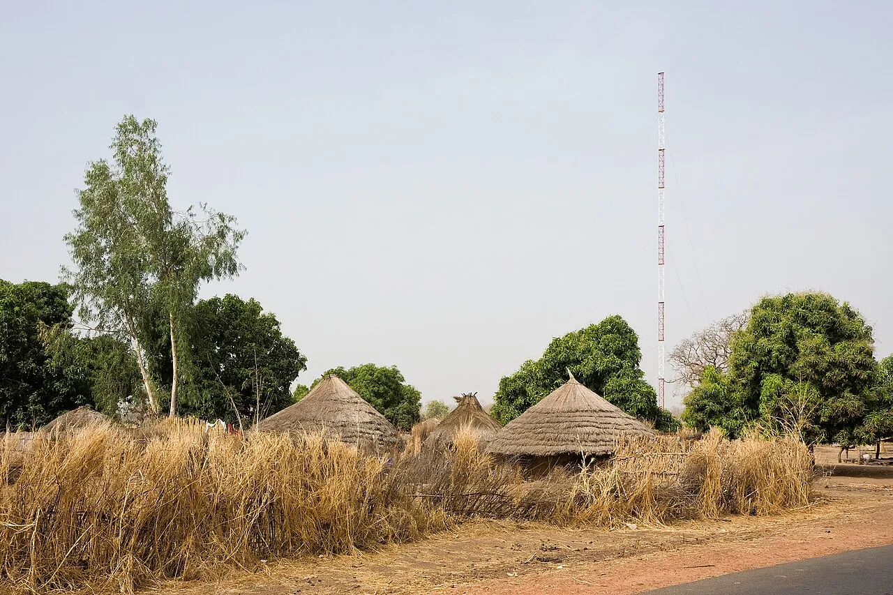 A mobile phone antenna rising above a rural village of thatched-roof huts in the Gambia.