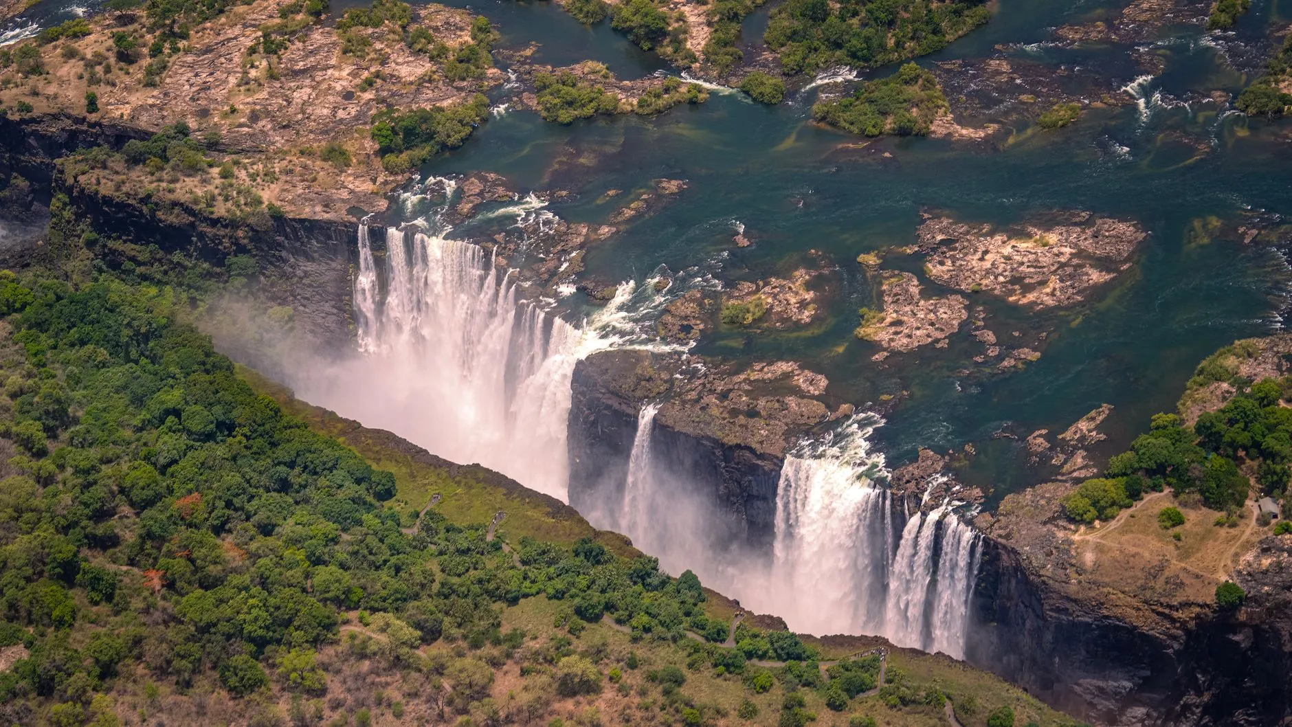 From the air, the Zambezi plunges into the narrow basalt gorge of Victoria Falls, framed by green bush.