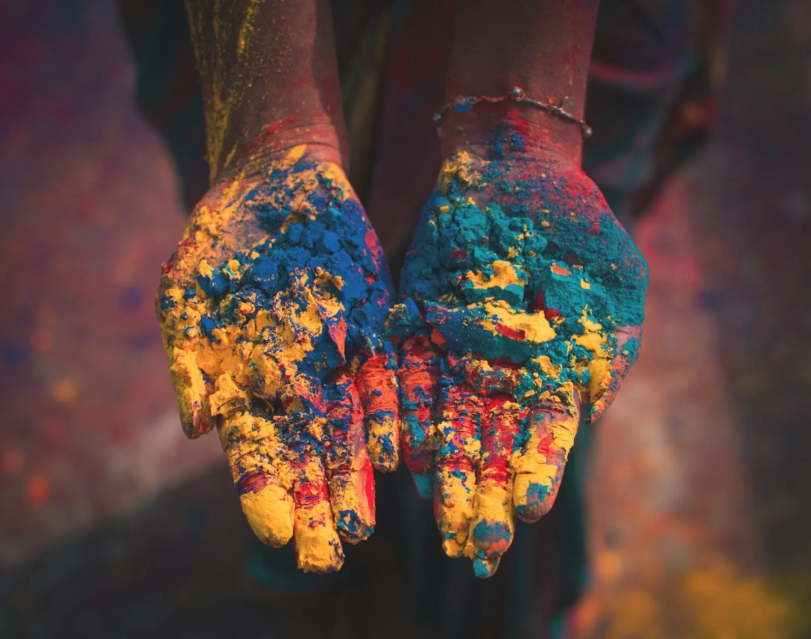 Two cupped hands held out and covered in yellow, blue and red coloured powder, against a blurred dark background