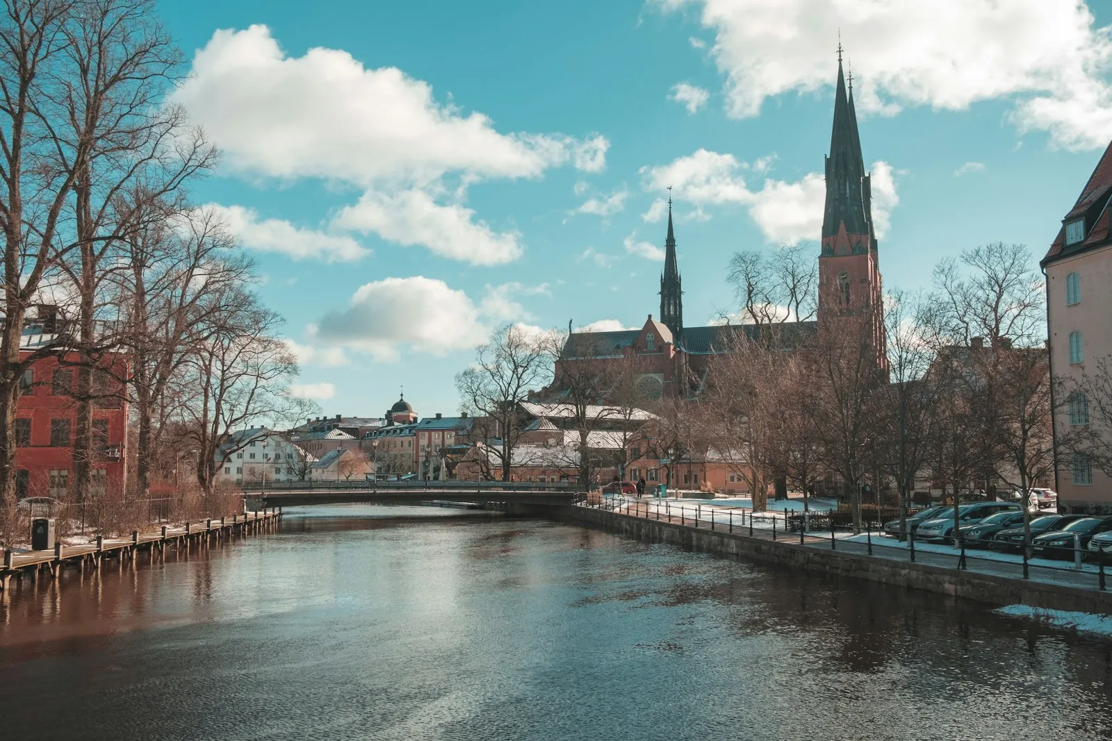 The twin spires of Uppsala Cathedral seen across the river from a bridge on a sunny late-winter day, with bare trees and patches of snow along the quay.