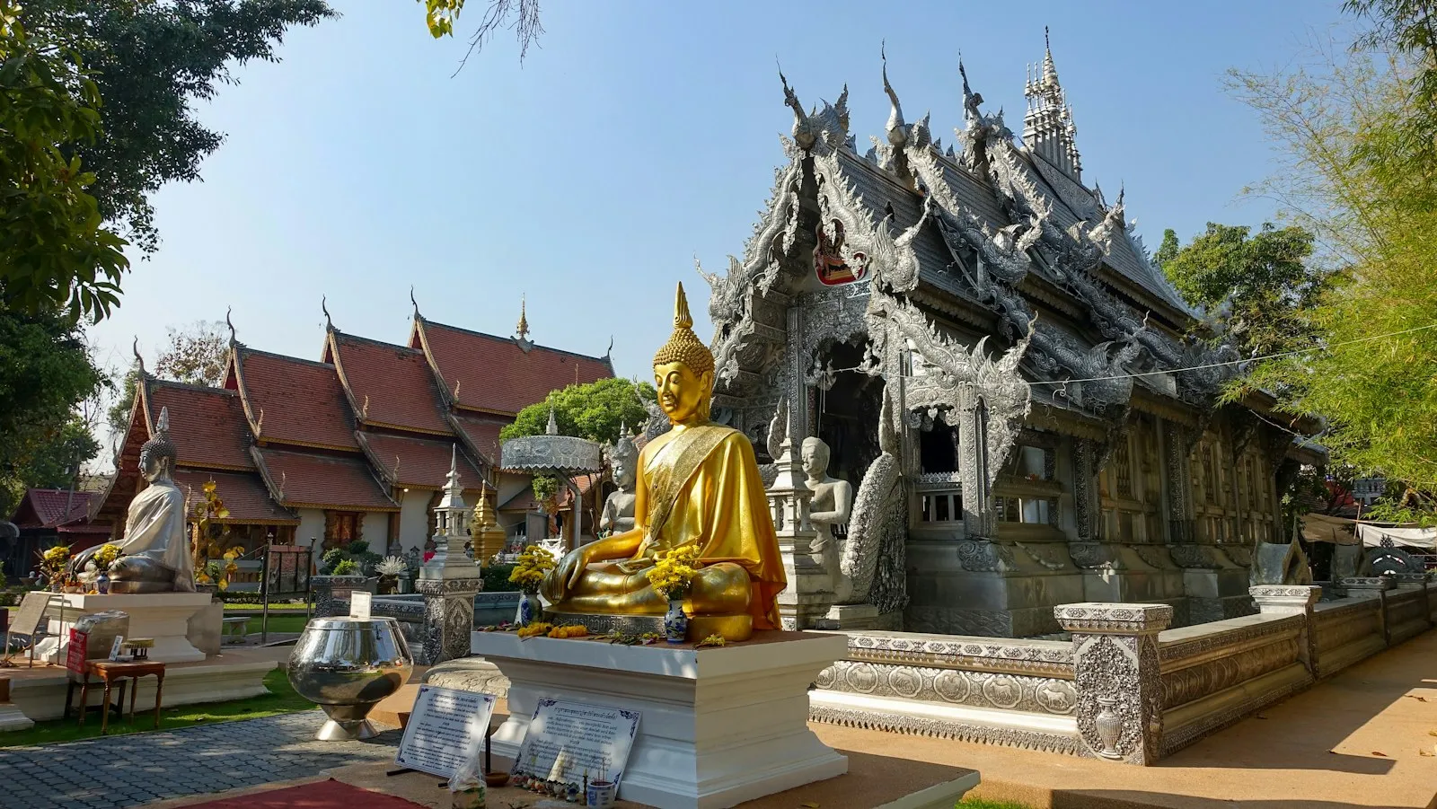 A golden seated Buddha in front of a silver-clad temple hall in Chiang Mai, with red-roofed temple buildings behind