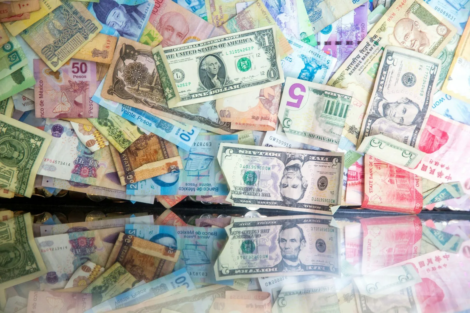 Banknotes from many countries spread out in a pile, with a one-dollar and a five-dollar US bill on top, mirrored in a glass surface