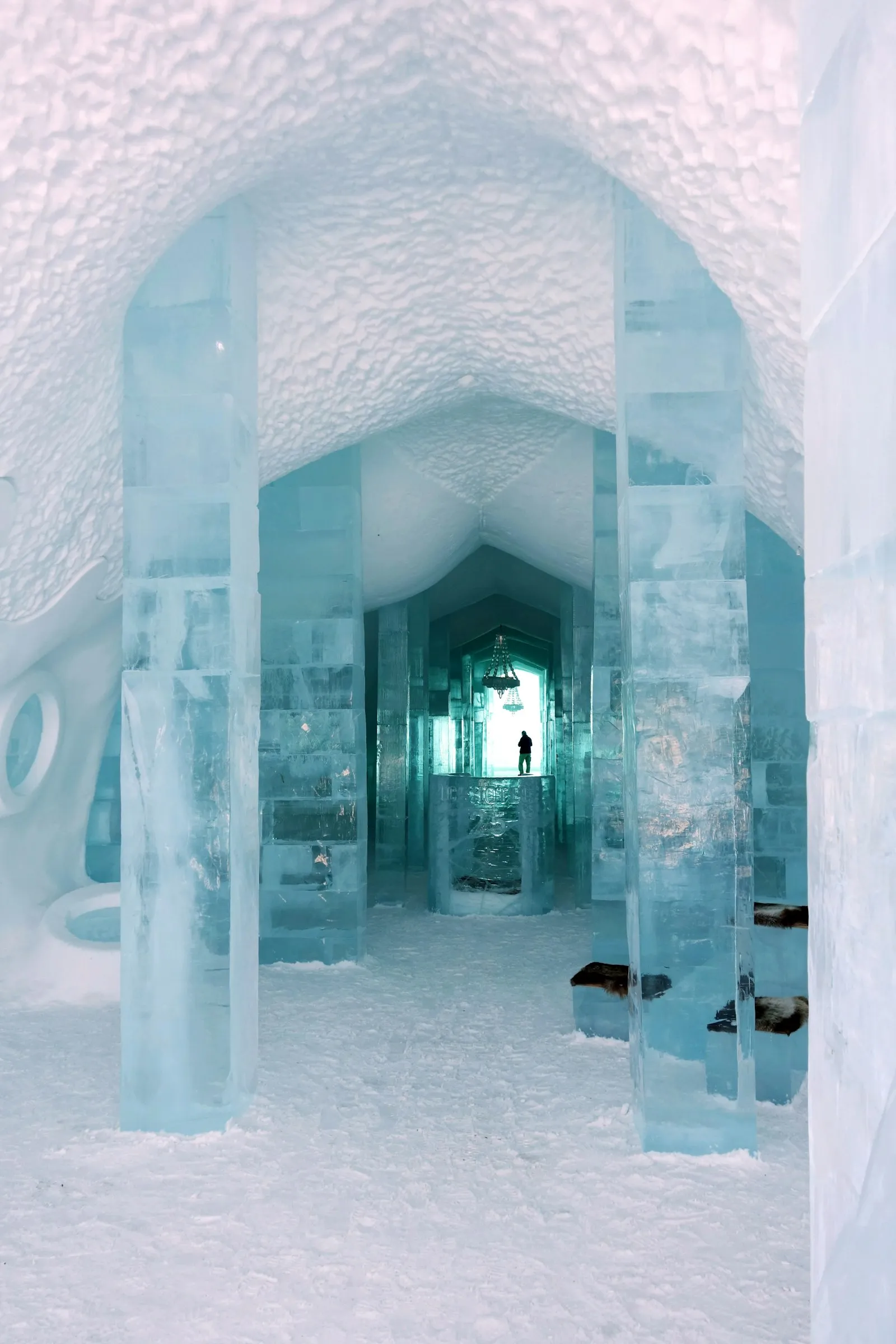 Hallway of the Icehotel with translucent ice pillars, a snow-vaulted ceiling and a lone visitor at the far end under an ice chandelier.