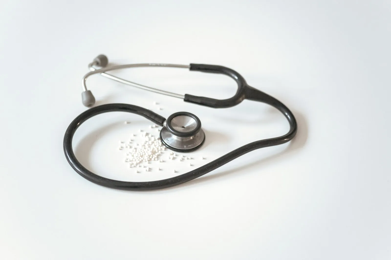 A black stethoscope with a steel chest piece lying in a loop on a white surface, with a small scatter of white pills inside the loop.
