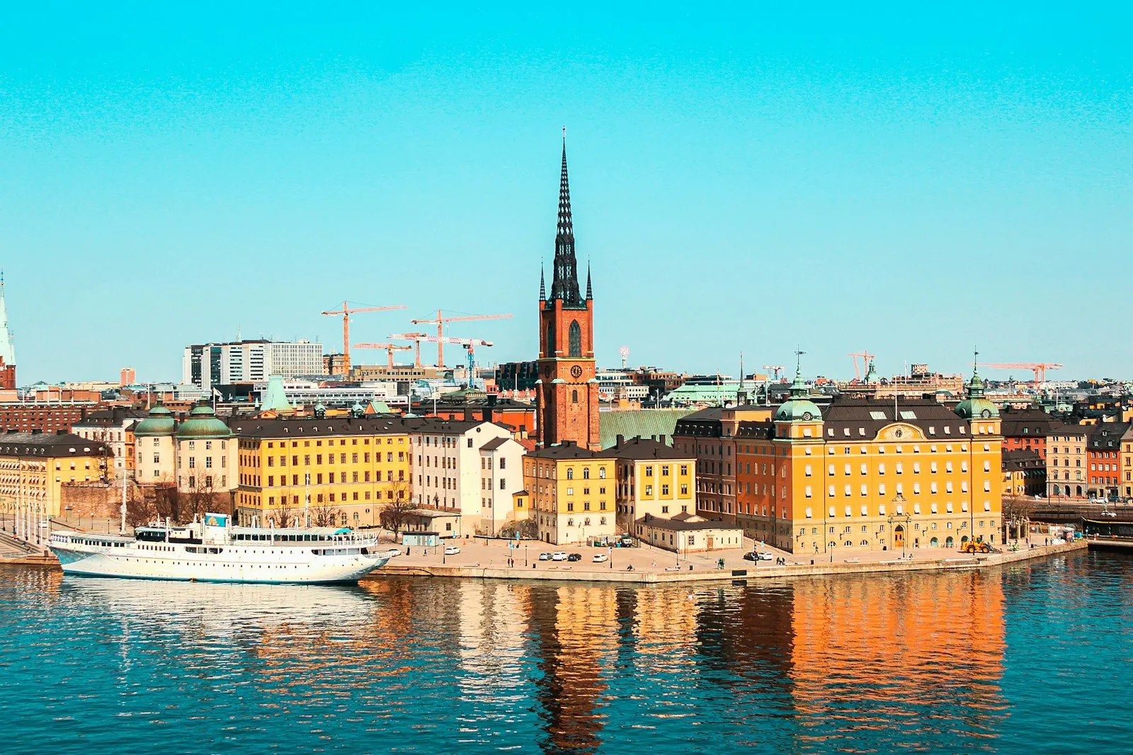Riddarholmen seen across the water, with the dark iron spire of Riddarholmen Church rising above ochre and yellow buildings and a white boat moored at the quay