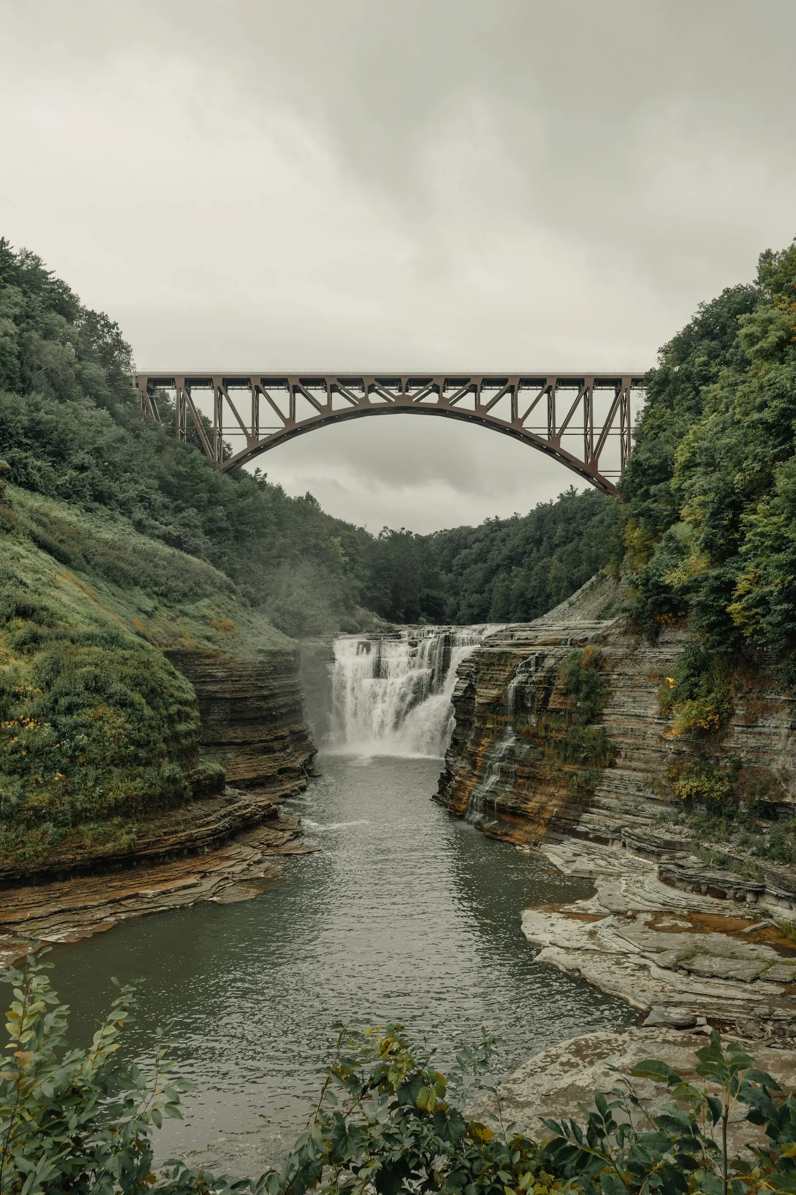 Waterfall dropping over layered rock ledges into a wooded gorge, with a steel arch railway bridge spanning the valley above under an overcast sky.
