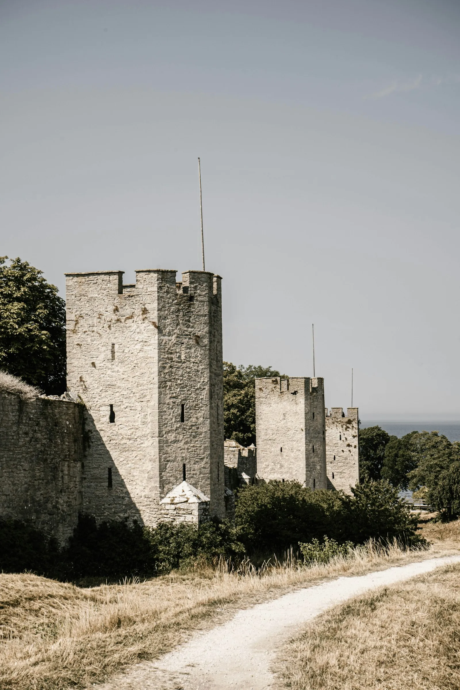 Three square limestone towers of Visby's ring wall above a gravel path and dry grass, with the Baltic Sea behind