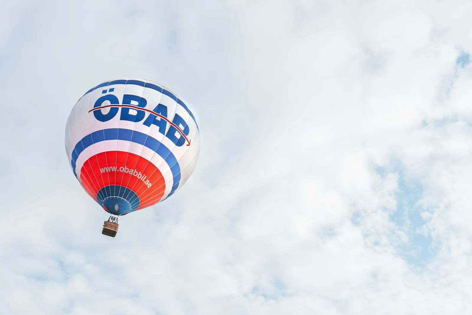 A red, white and blue hot-air balloon with ÖBAB lettering drifting under a pale, cloudy sky.