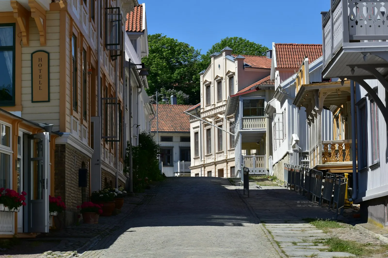A cobbled, car-free street in Marstrand lined with wooden houses with carved balconies and a Hotell sign, under a clear blue sky.
