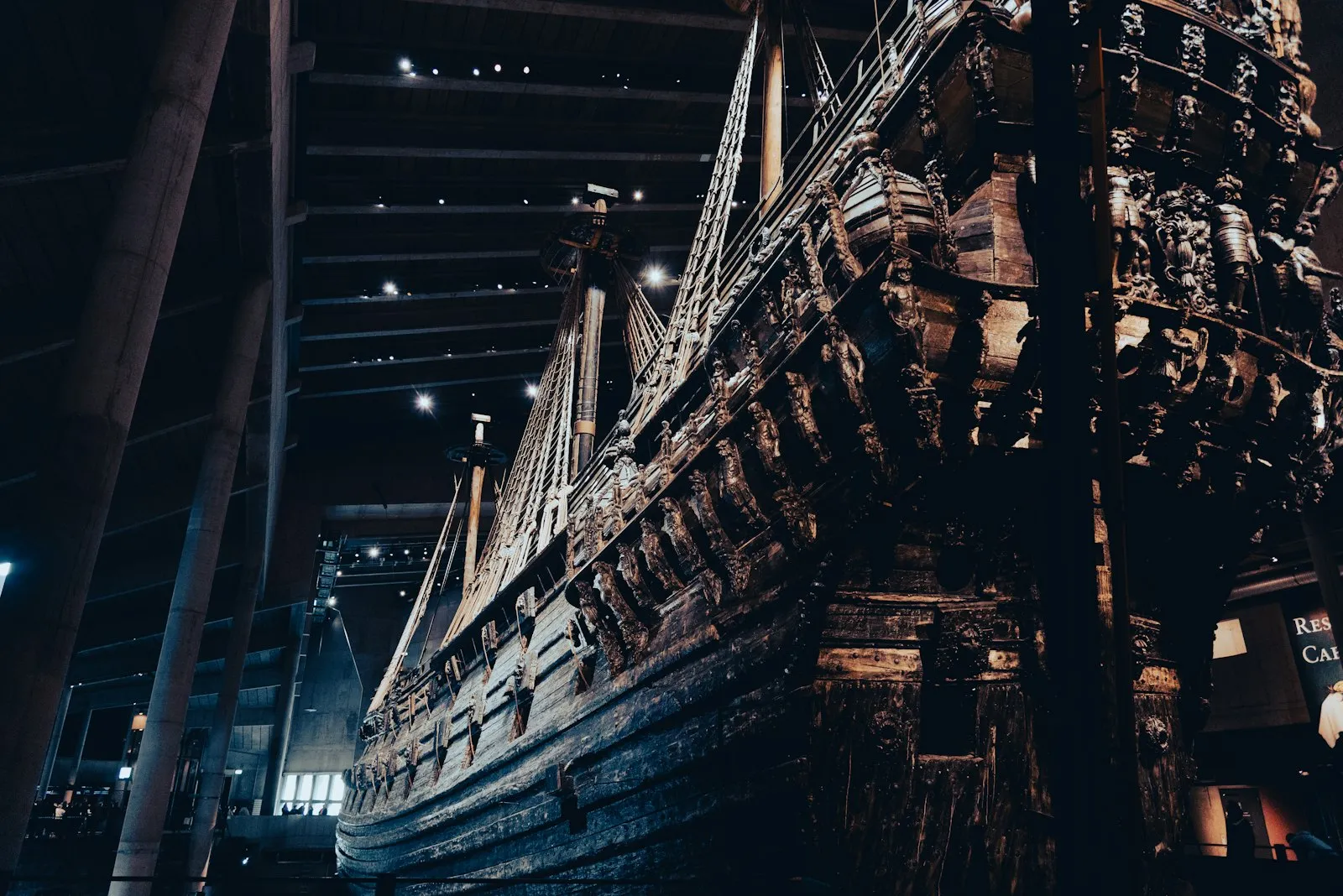 The dark carved hull and masts of the warship Vasa seen from below inside the dimly lit museum hall, with concrete pillars and ceiling lights behind
