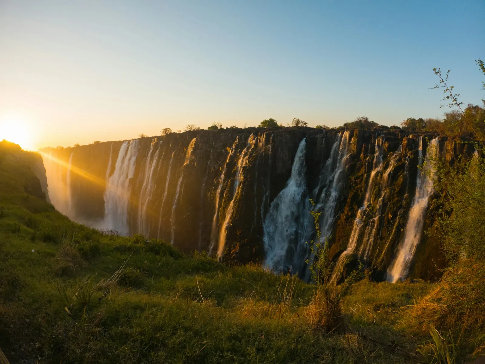 Victoria Falls seen from the Zambian side at low sun, water pouring over the long basalt cliff edge.