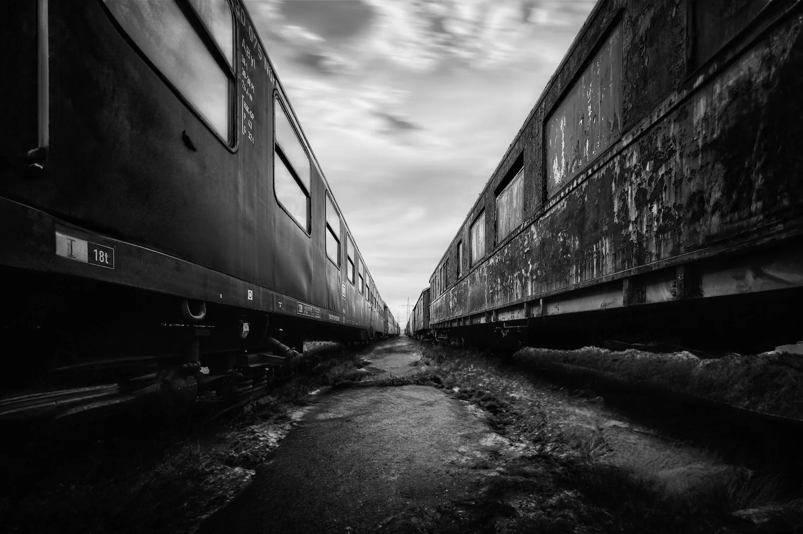 Black-and-white view down a narrow gap between two weathered railway carriages on a siding, rust and peeling paint on the coaches, cloudy sky above.