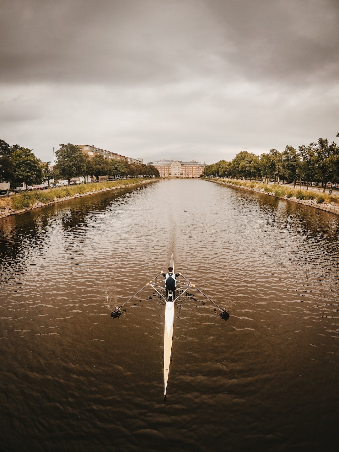 A single rower in a narrow racing scull on a wide, tree-lined canal in Malmö under heavy grey cloud, with a long brick building closing the view.