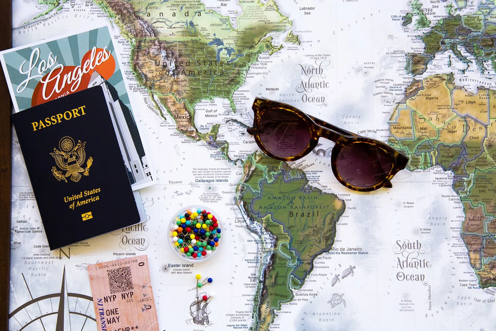 A United States passport, a Los Angeles guidebook, tortoiseshell sunglasses, a train ticket and a jar of coloured map pins laid out on a world map of the Americas and the Atlantic.