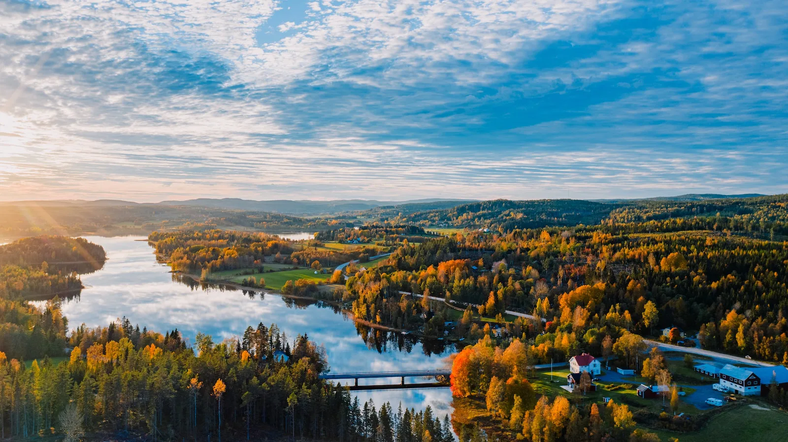 Aerial view of a long lake with a road bridge, autumn-coloured birch and pine forest, a few red and white farmhouses and low forested hills under a partly cloudy sky at sunrise.