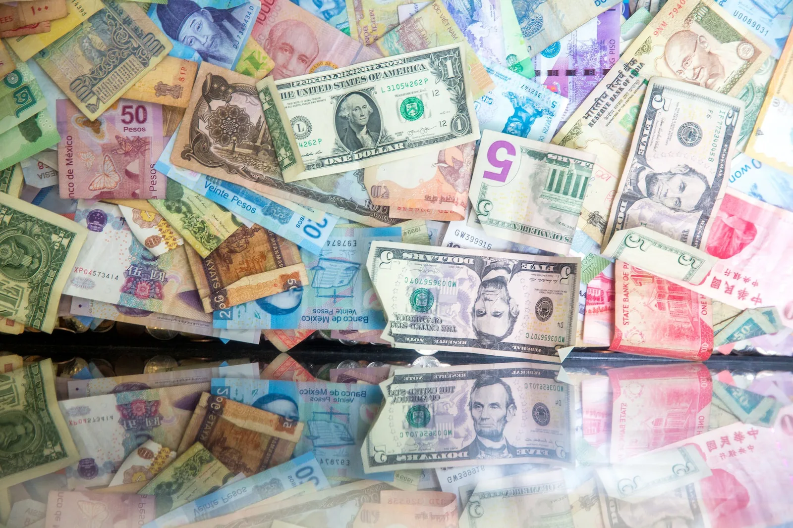 Banknotes from many countries spread out in a pile, with a one-dollar and a five-dollar US bill on top, mirrored in a glass surface