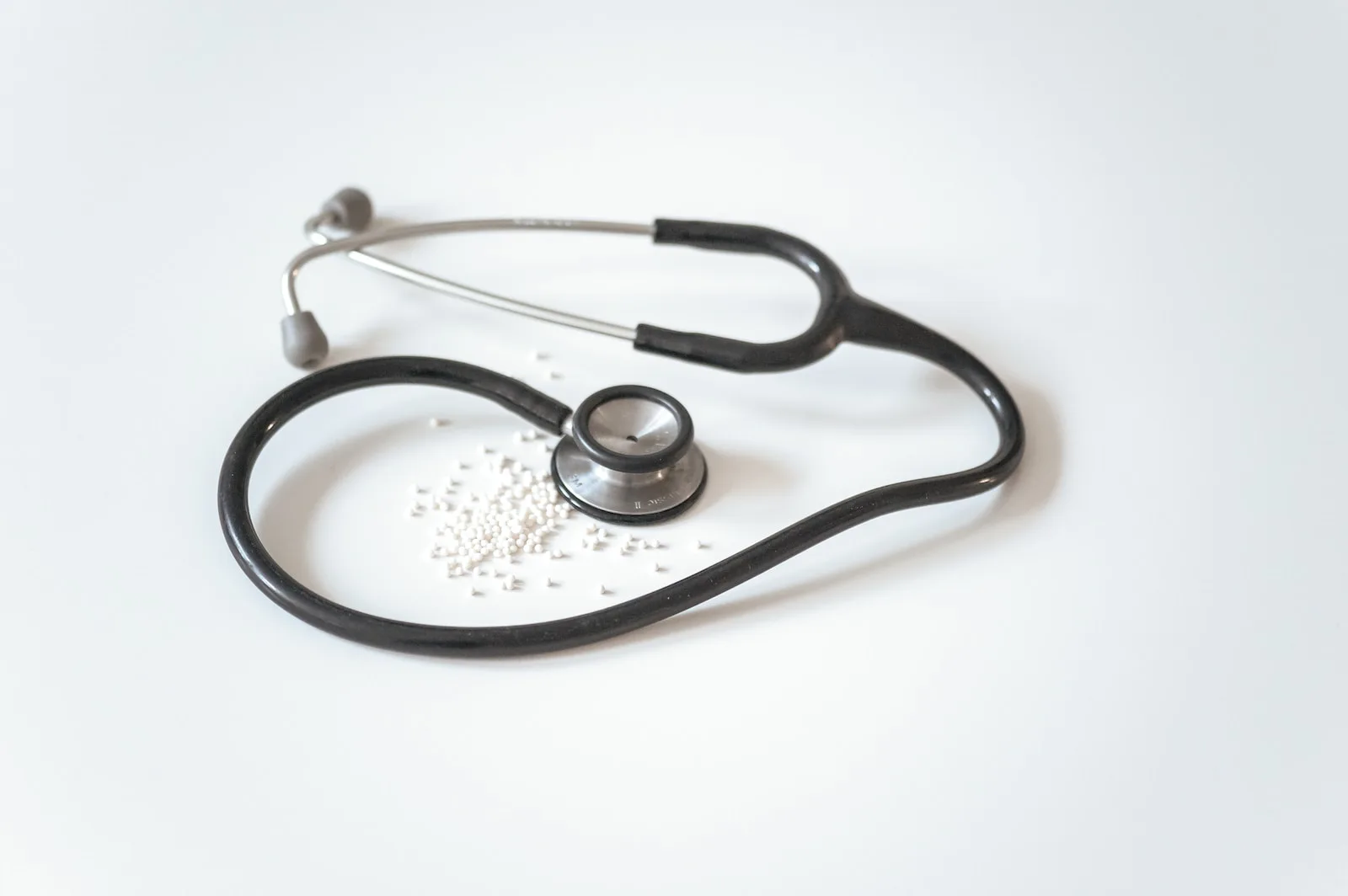 A black stethoscope with a steel chest piece lying in a loop on a white surface, with a small scatter of white pills inside the loop.