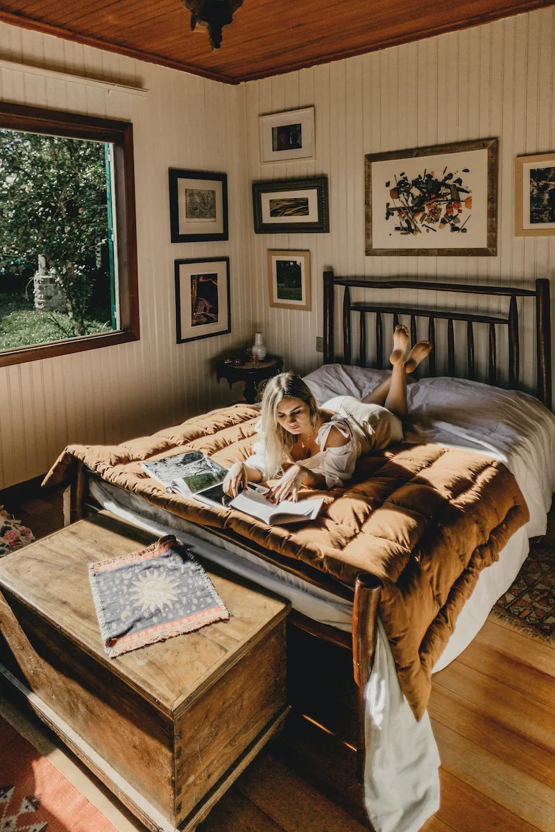 Sunlit wood-panelled cabin bedroom with an open window onto green trees, a woman lying on a bed under a brown quilt reading a book, framed pictures on the wall and a wooden chest at the foot of the bed.